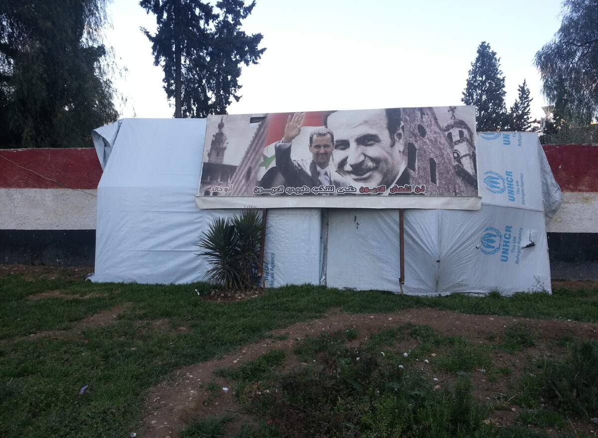 Reem Alghazzi, photographie tirée du Livre noir des Assad, représentant l’entrée d’une tente de réfugiés de l’UNHCR surplombée par une affiche de Bachar et Hafez al-Assad. C Reem Alghazzi.